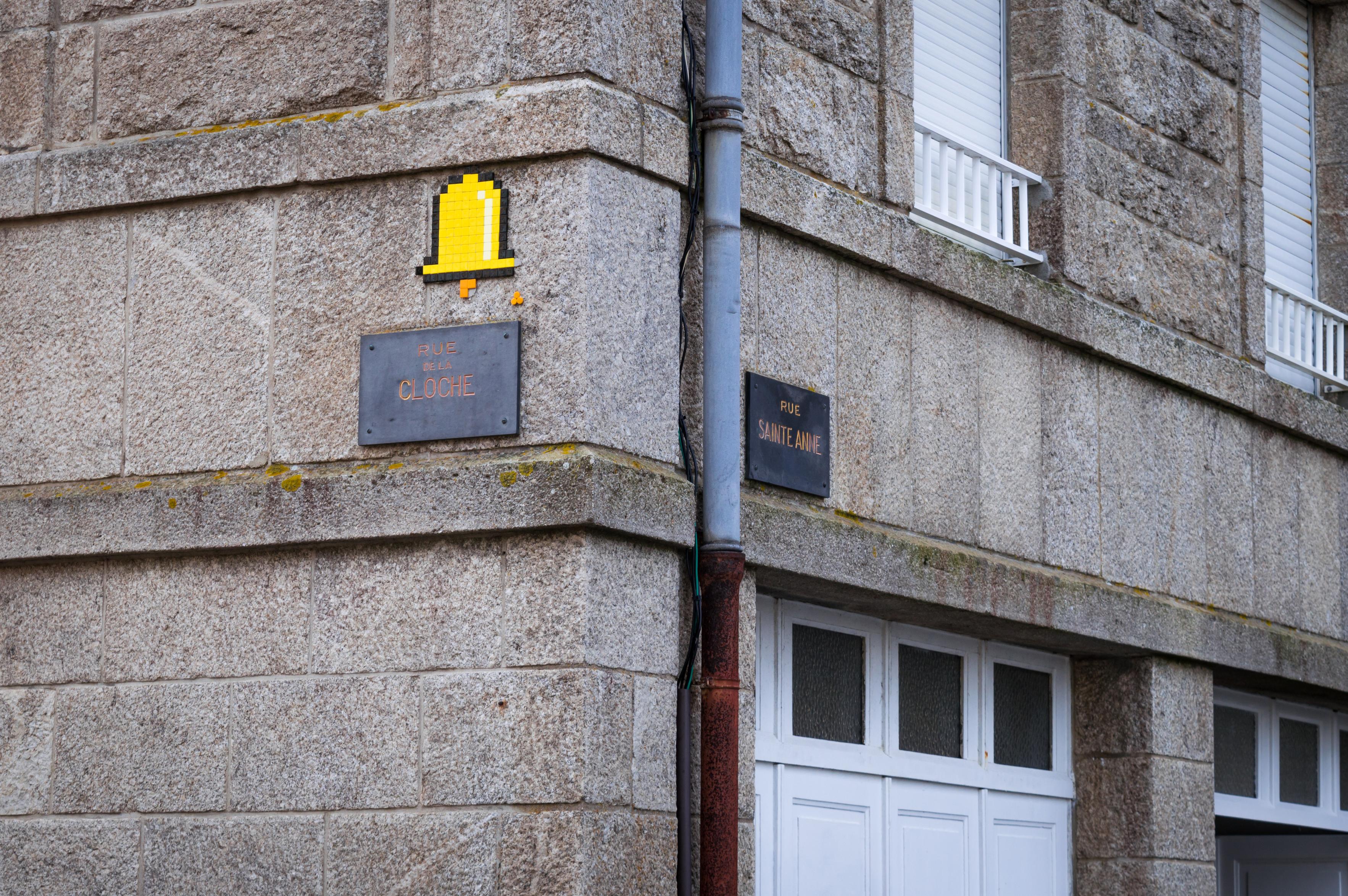 Wall of a bulding. One side has a sign reading "Rue Sainte Anne" and the other – "Rue de la Cloche" (Bell Street). Small pixelart-style graffiti of a yellow bell has been placed above it.