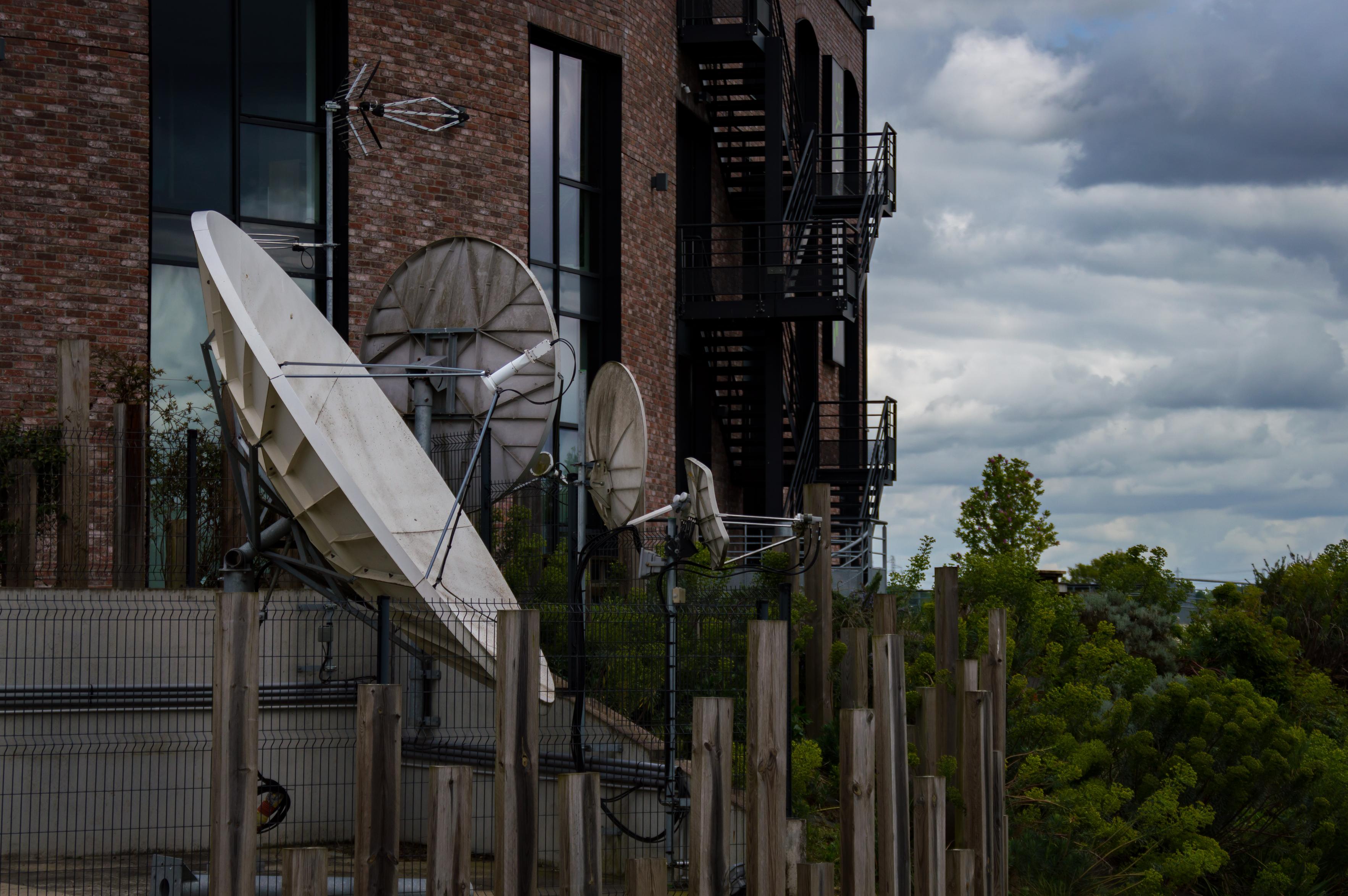 A set of various dish antennas and other radio communications equipment set up in a rig next to a red brick building. Greenery and clouds can be seen in the right part of the frame